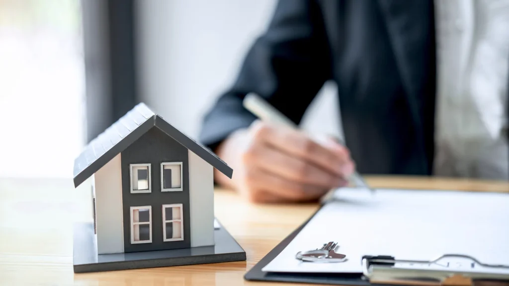 Benefits of a Financial Plan - Man signing a document on a clipboard with a wooden, model house sitting on the table in the foreground