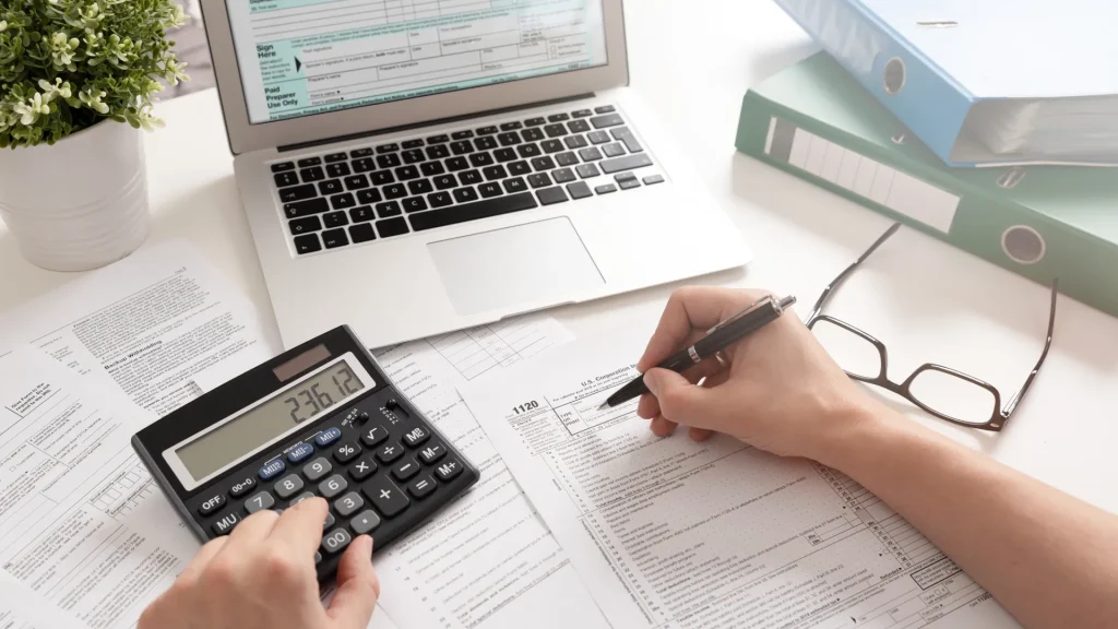 Person preparing for 2025 tax deadlines by filling out documents with pen, calculator, and laptop